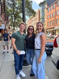 Three young adults smiling on a sunny downtown sidewalk by a lamppost, wearing casual summer outfits with brick storefronts, trees and parked cars in the urban neighborhood