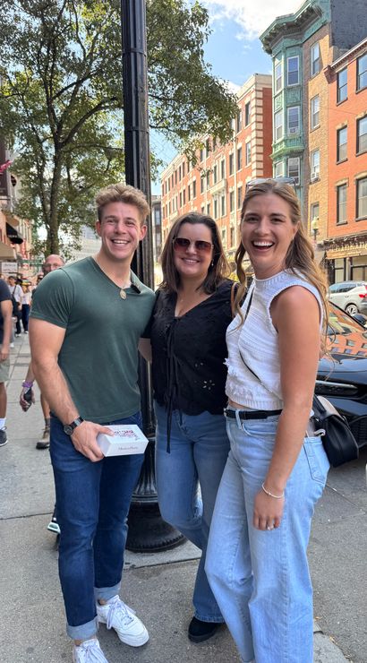 Three young adults smiling on a sunny downtown sidewalk by a lamppost, wearing casual summer outfits with brick storefronts, trees and parked cars in the urban neighborhood