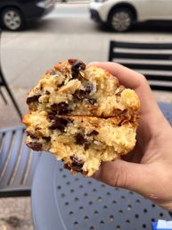 Close-up of a hand holding a halved chocolate-chip muffin with gooey melted chocolate, outdoor cafe table and parked cars on a city sidewalk in the background.