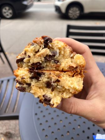 Close-up of a hand holding a halved chocolate-chip muffin with gooey melted chocolate, outdoor cafe table and parked cars on a city sidewalk in the background.