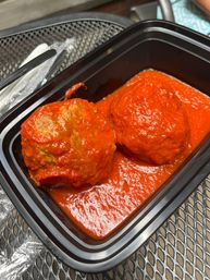 Close-up of two large meatballs smothered in bright red marinara sauce inside a black takeout container on a metal patio table.