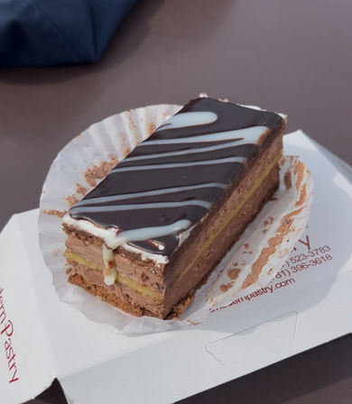 Chocolate layered dessert bar with glossy dark ganache and white icing drizzle, sitting in a paper liner inside a takeout box on a café table.