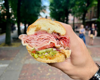 Close-up of a handheld deli sandwich with thinly sliced ham and salami, tomato, lettuce, pickles and red onion on a crusty roll, held over a tree-lined city sidewalk