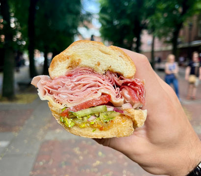 Close-up of a handheld deli sandwich with thinly sliced ham and salami, tomato, lettuce, pickles and red onion on a crusty roll, held over a tree-lined city sidewalk