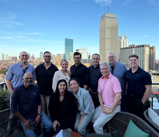 Casual rooftop gathering of adults smiling on a patio with the Boston skyline and skyscrapers under a clear blue sky.