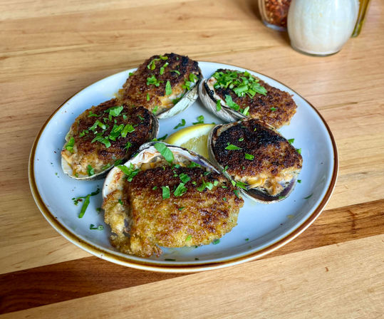 Golden-brown baked stuffed clams on half shells with crispy breadcrumb topping, chopped parsley and lemon wedge on a white plate set on a wooden table, seafood appetizer.