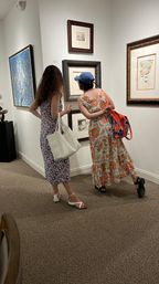 Two visitors in floral sundresses view framed drawings and paintings on white gallery walls in a carpeted art gallery interior; one carries a white tote, the other wears a blue cap and orange bag — exhibition scene.