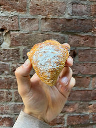 Hand holding a golden, flaky sfogliatella pastry dusted with powdered sugar against a weathered red brick wall backdrop