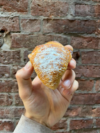 Hand holding a golden, flaky sfogliatella pastry dusted with powdered sugar against a weathered red brick wall backdrop