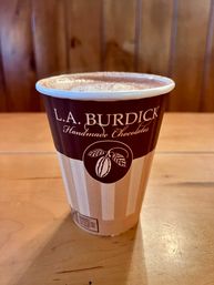 Rich frothy hot chocolate in a striped brown paper cup on a wooden table with a warm wood-paneled café backdrop.