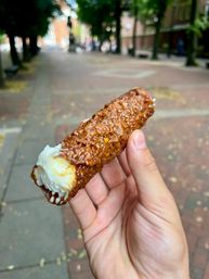 Hand holding a crunchy caramel-and-nut coated pastry tube filled with swirled vanilla cream, street snack photographed on a tree-lined city sidewalk.