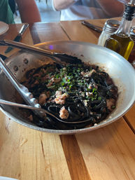 Skillet of squid-ink pasta with chopped seafood and fresh herbs, served with tongs on a wooden table next to olive oil and a salt shaker — rustic Italian-style seafood pasta.