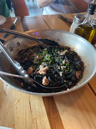Skillet of squid-ink pasta with chopped seafood and fresh herbs, served with tongs on a wooden table next to olive oil and a salt shaker — rustic Italian-style seafood pasta.