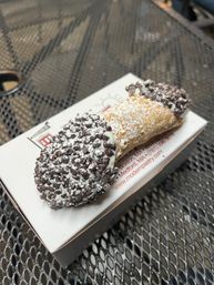 Chocolate-chip cannoli dusted with powdered sugar on a pastry box, sitting on a metal outdoor cafe table — crispy shell filled with creamy ricotta-style filling.