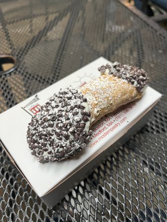 Chocolate-chip cannoli dusted with powdered sugar on a pastry box, sitting on a metal outdoor cafe table — crispy shell filled with creamy ricotta-style filling.
