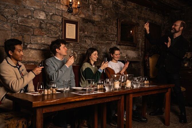 Four people seated at a wooden table enjoying a guided whisky tasting in a cozy, candlelit stone-walled cellar as an instructor holds up a glass.