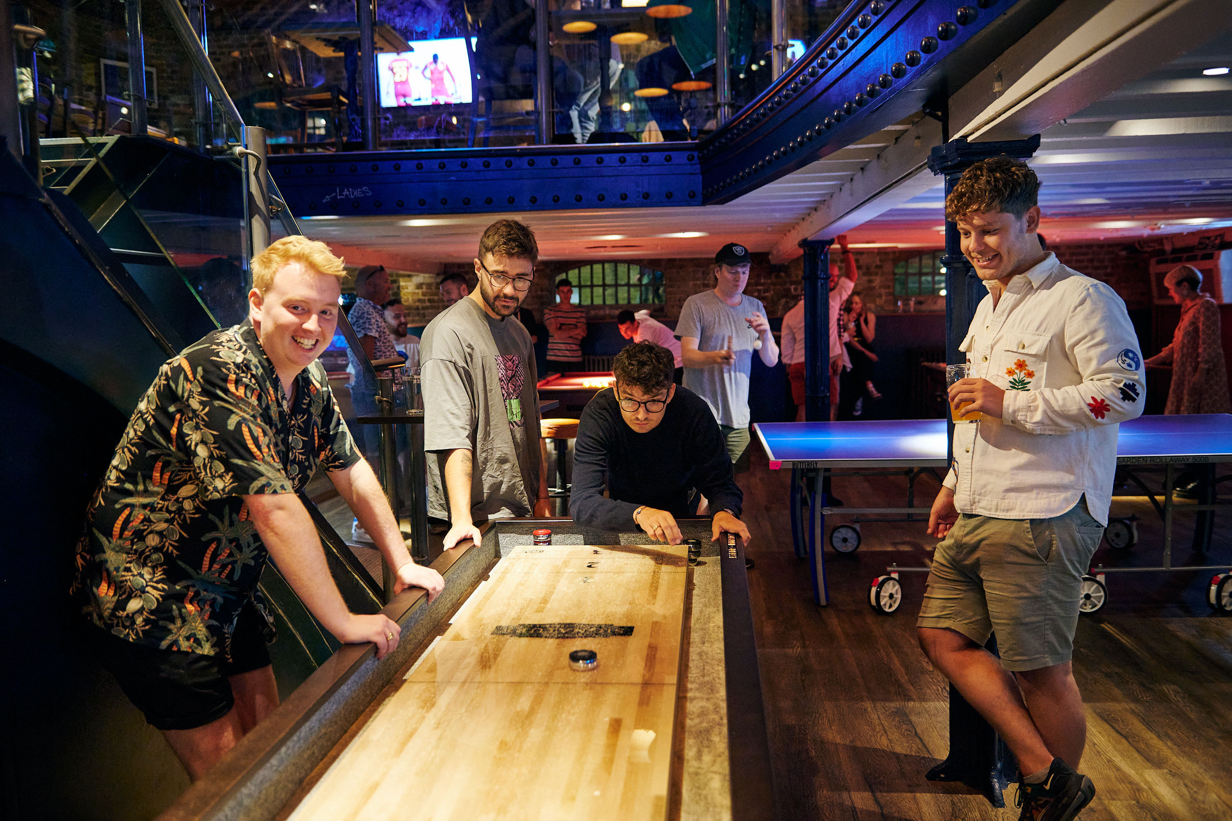 Friends laughing around a shuffleboard table in a lively indoor bar with ping-pong tables and drinks