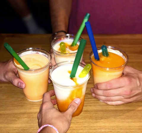 Four frozen mango-and-cream cocktails in clear plastic cups with colorful straws and tiny pipette droppers, held together in a cheerful toast over a wooden table — summer bar vibes.