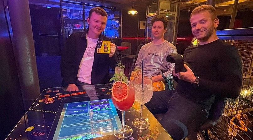 Three people at an arcade bar table with a built-in touchscreen game in a neon-lit lounge, two colorful cocktails with citrus garnish on the glass surface, one person holding a retro game controller and another holding a novelty mug — upbeat nightlife scene.