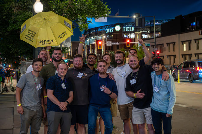 Group of young men smiling on a Denver night out near a lit stadium, posing under a yellow umbrella during a pub crawl