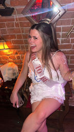 Laughing woman in a white floral dress wearing a 'Bride to Be' sash at a dim, brick-walled bar, holding a drink and phone with festive bachelorette party vibes.