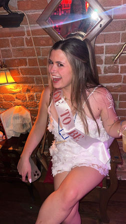 Laughing woman in a white floral dress wearing a 'Bride to Be' sash at a dim, brick-walled bar, holding a drink and phone with festive bachelorette party vibes.