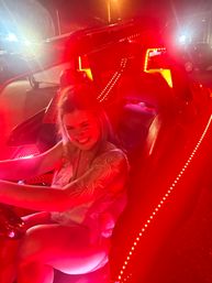 Smiling person behind the wheel of an LED-lit convertible at night, vivid red interior lights and parking-lot neon glow
