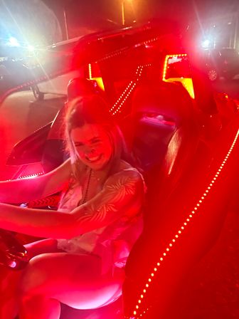 Smiling person behind the wheel of an LED-lit convertible at night, vivid red interior lights and parking-lot neon glow