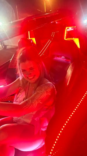 Smiling person behind the wheel of an LED-lit convertible at night, vivid red interior lights and parking-lot neon glow