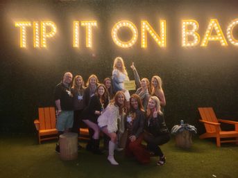 Group of friends posing beneath large illuminated marquee letters reading 'TIP IT ON BACK' at a turf lounge, orange Adirondack chairs and a small 'Denver' sign visible.