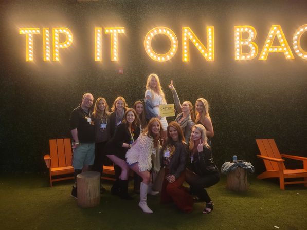Group of friends posing beneath large illuminated marquee letters reading 'TIP IT ON BACK' at a turf lounge, orange Adirondack chairs and a small 'Denver' sign visible.