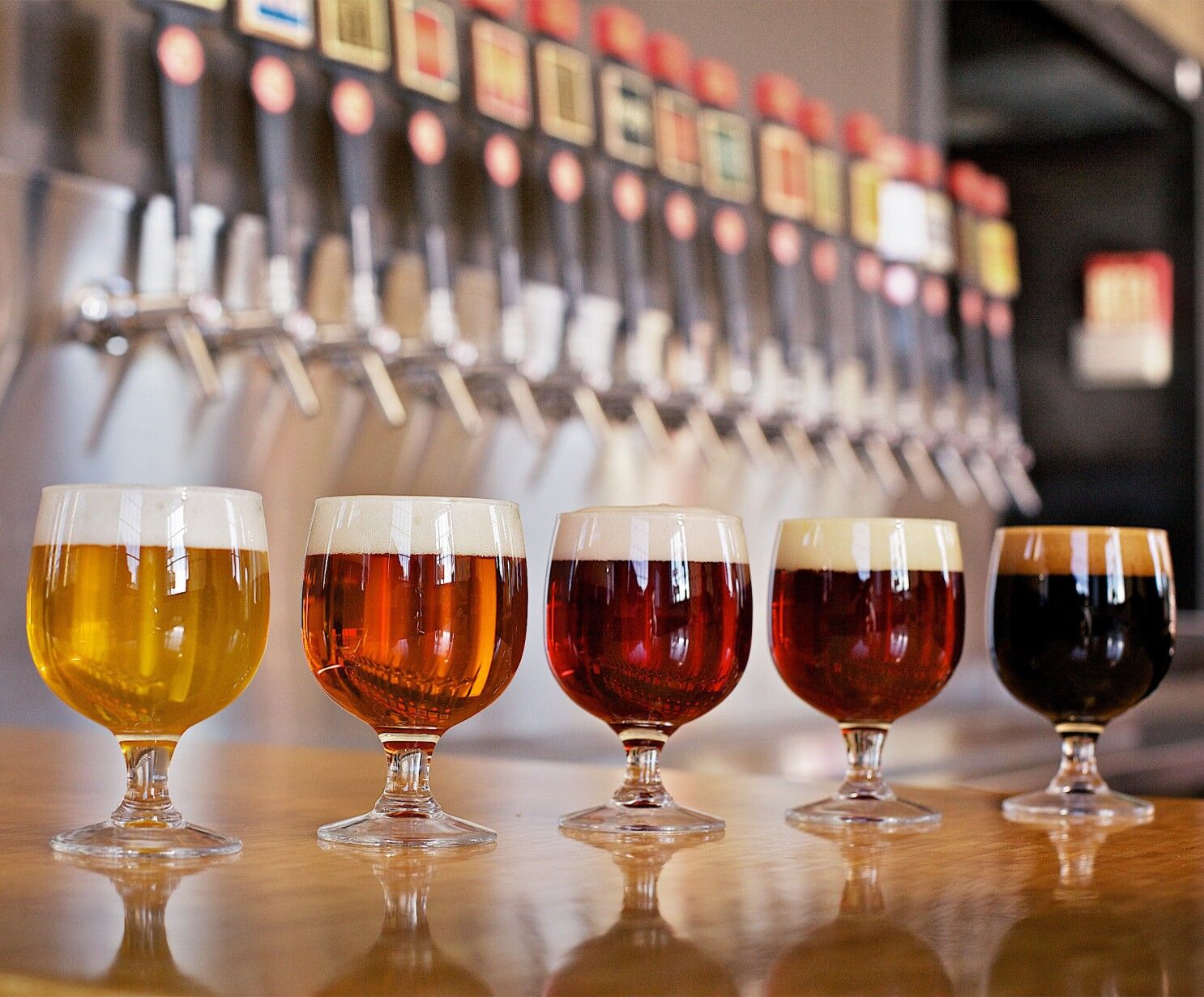 Colorful craft beer flight: five snifter glasses from pale gold to dark stout on a wooden bar with draft taps blurred in the background.