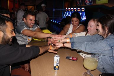 Friends at a lively bar-arcade doing a team cheer with hands stacked over a table, a Corona can and cocktail nearby and neon-lit skee-ball machines in the background.
