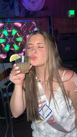 Young woman in white dress puckering a kiss while holding a lemon-garnished cocktail at a neon-lit bar with colorful projection and party sash