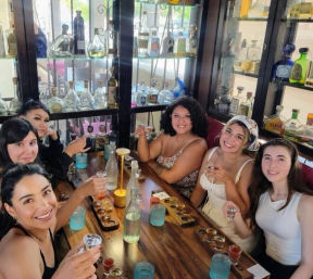 Six women smiling and raising shot glasses during a tequila tasting at a wooden table, blue tumblers and shelves of tequila bottles displayed behind them in a lively bar setting.