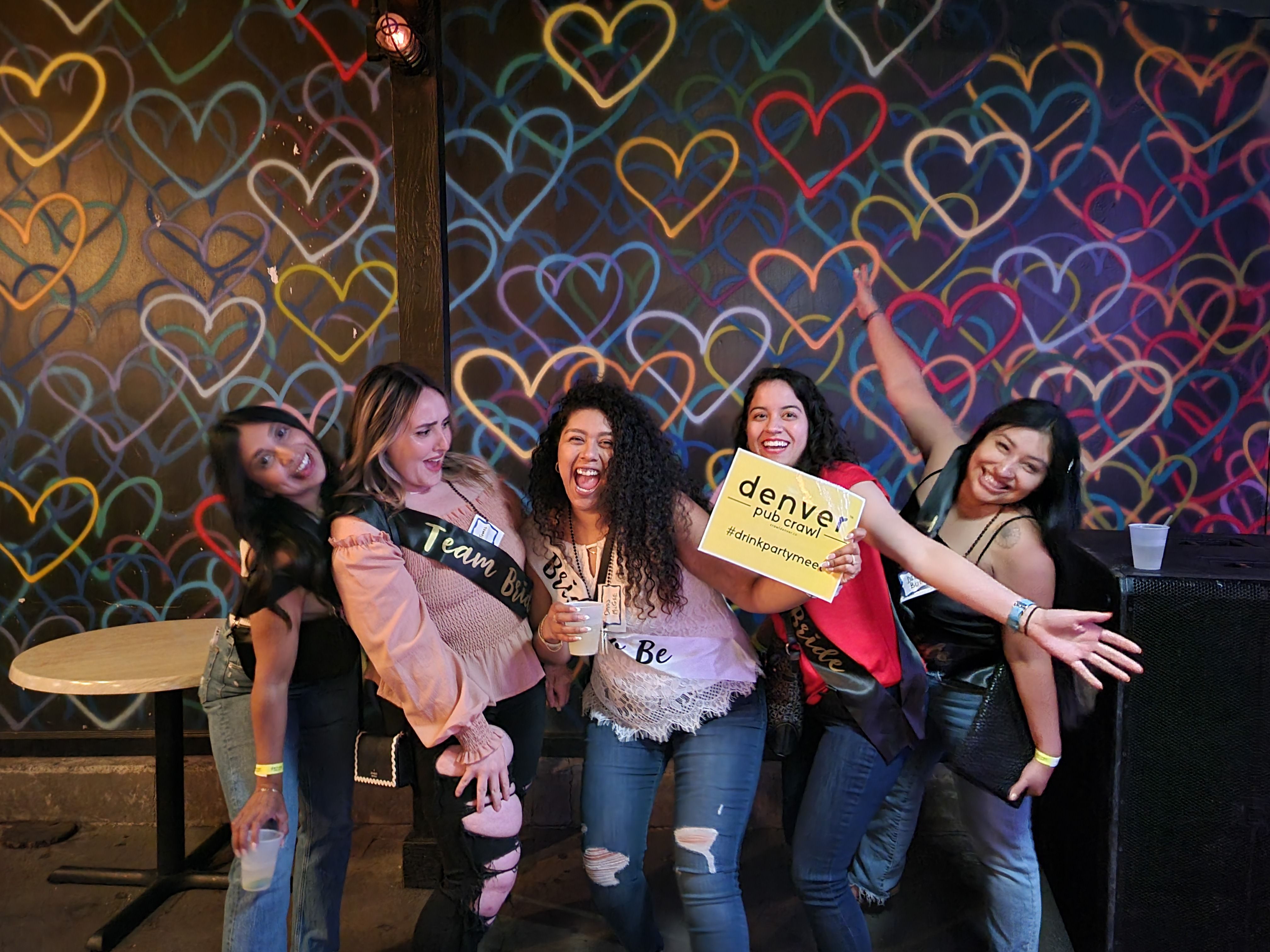 Five women celebrating a bachelorette party in a lively bar, wearing sashes and holding drinks and a sign while posing in front of a colorful neon heart mural.