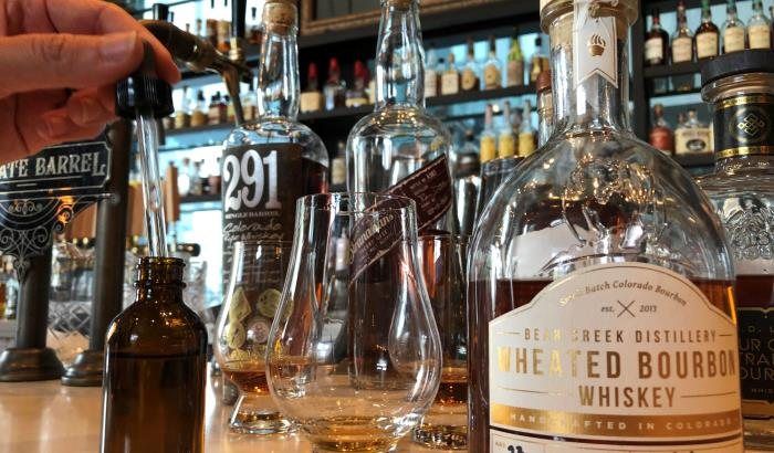 Close-up of a whiskey tasting on a bar: a hand uses a dropper into a small amber vial beside two Glencairn glasses with amber bourbon and assorted whiskey bottles on the shelf.