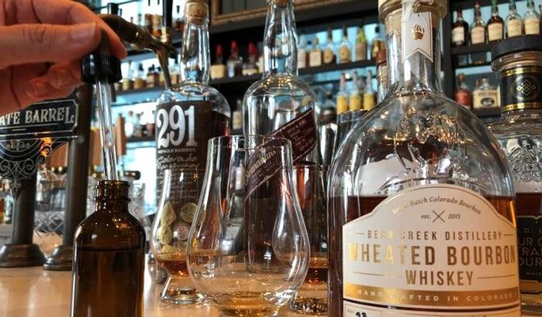 Close-up of a whiskey tasting on a bar: a hand uses a dropper into a small amber vial beside two Glencairn glasses with amber bourbon and assorted whiskey bottles on the shelf.