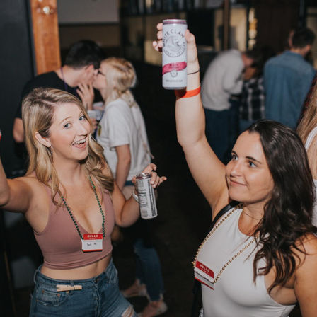 Two young women at an indoor bar raising canned drinks and smiling, festive nightlife scene with people mingling in the background.