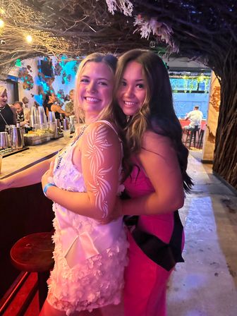 Two smiling women hugging at a lively bar—one in a white floral dress with white arm paint, the other in a pink dress—under twinkling string lights and a rustic branch canopy.