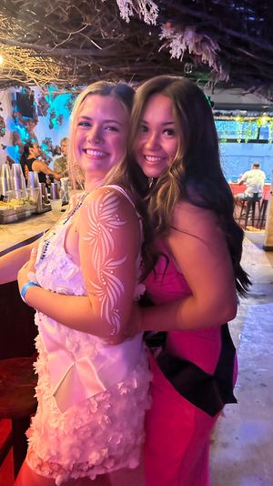 Two smiling women hugging at a lively bar—one in a white floral dress with white arm paint, the other in a pink dress—under twinkling string lights and a rustic branch canopy.