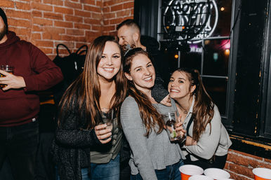 Three smiling young women holding cocktails in a brick‑walled urban bar with a neon window sign and red solo cups on a table, lively nightlife scene