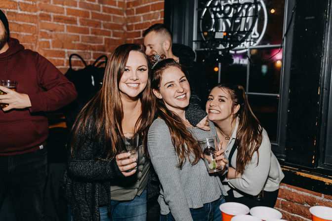 Three smiling young women holding cocktails in a brick‑walled urban bar with a neon window sign and red solo cups on a table, lively nightlife scene