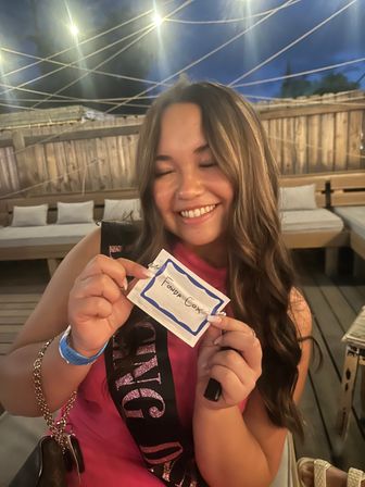 Smiling woman in a pink dress wearing a glittery bachelorette sash, holding a handwritten name tag on a rooftop patio at night under crisscrossed string lights and wooden fencing.