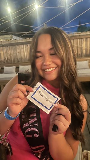 Smiling woman in a pink dress wearing a glittery bachelorette sash, holding a handwritten name tag on a rooftop patio at night under crisscrossed string lights and wooden fencing.