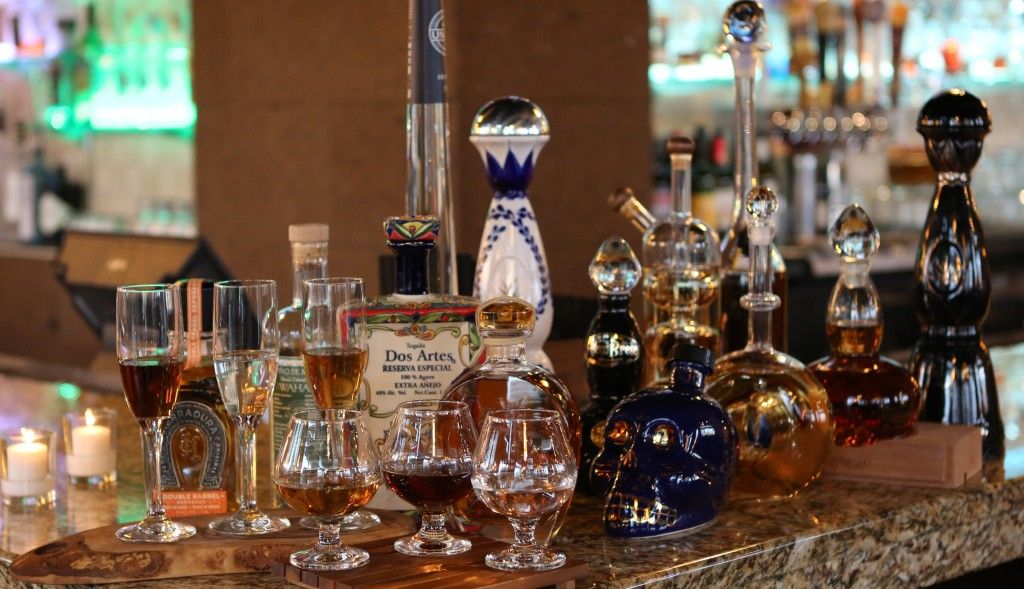Assorted premium tequila and spirits in decorative bottles and decanters, including a skull-shaped bottle, with tasting snifters and stemmed glasses on a marble bar counter, lit candles and warm lounge lighting in the background.