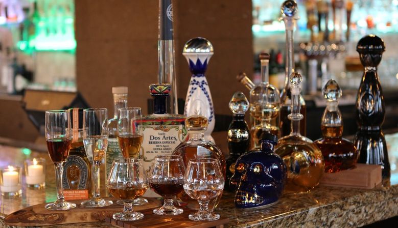 Assorted premium tequila and spirits in decorative bottles and decanters, including a skull-shaped bottle, with tasting snifters and stemmed glasses on a marble bar counter, lit candles and warm lounge lighting in the background.