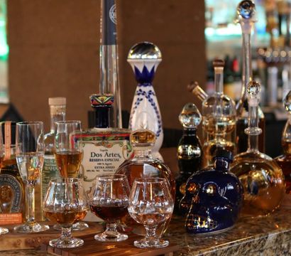 Assorted premium tequila and spirits in decorative bottles and decanters, including a skull-shaped bottle, with tasting snifters and stemmed glasses on a marble bar counter, lit candles and warm lounge lighting in the background.