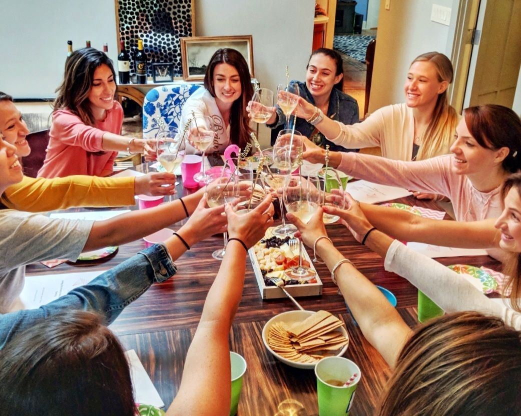 Cheerful group of women toasting with wine glasses around an indoor dining table with a charcuterie board and crackers at a casual at-home gathering.