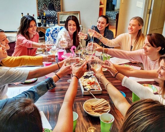 Cheerful group of women toasting with wine glasses around an indoor dining table with a charcuterie board and crackers at a casual at-home gathering.